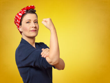 A woman posing like Rosie the Riveter flexing her arm on a yellow background