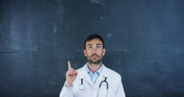 A medical professor in white coat stands in front of a chalkboard pointing up.
