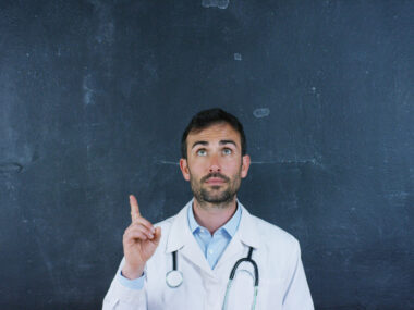 A medical professor in white coat stands in front of a chalkboard pointing up.