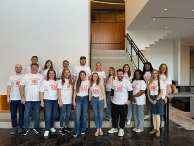 Eighteen people all wearing the same white T-shirt with a red logo pose for a group photo in a large room in front of a set of stairs.