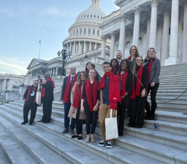 Thirteen people with various red items of clothing, such as scarves, stand on the steps of Capitol Hill in Washington, D.C. for a group photo.