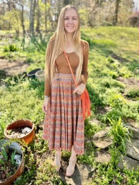 A woman with long blond hair wears a brown top and a skirt amid a clearing of greenery, with trees not far away.