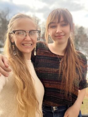 In an outdoor setting, a woman with long blond hair, long earrings, and glasses stands beside a younger woman with long brown hair. The younger woman's arm is around the first woman's shoulder.