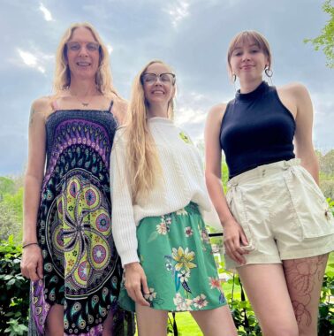 A photo taken from a low angle features three women standing side by side and smiling. They are outdoors, and it looks to be a warm spring or summer day.