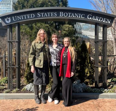 Three women stand side by side under an arched sign that reads "United States Botanic Garden." It's sunny, but their clothes suggest it's chilly outside. All are smiling straight ahead at the camera.