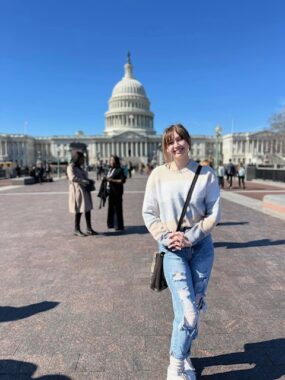A young woman in somewhat ripped jeans and a light top wears a shoulder bag as she stands on asphalt before the Capitol in Washington, D.C.