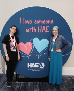 Two women stand in front of a large, blue sign in the shape of a circle. In pink writing, the sign reads "I love someone with HAE." At the bottom is the HAEA logo. 