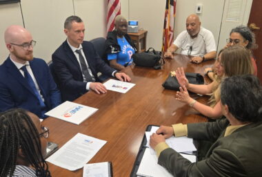 A group of people sit around a rectangular table in a small meeting room. There is an American flag and a Maryland flag in the far corner.