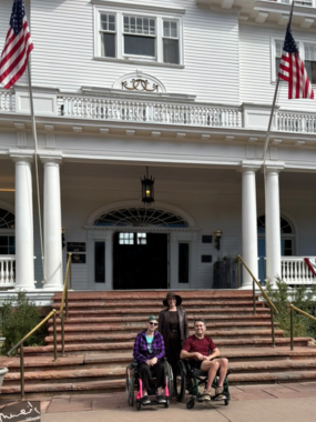 Two people in wheelchairs and one woman standing behind them pose in front of a large staircase and the entrance of a large, white, historic building with two U.S. flags flying on either side. 